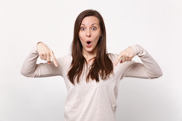 Amazed shocked young woman in light clothes keeping mouth open, pointing index fingers down isolated on white wall background in studio. People sincere emotions, lifestyle concept. Mock up copy space.