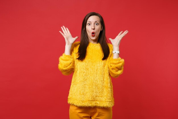 Amazed shocked young woman in yellow fur sweater keeping mouth open, spreading hands isolated on bright red wall background in studio. People sincere emotions, lifestyle concept. Mock up copy space.