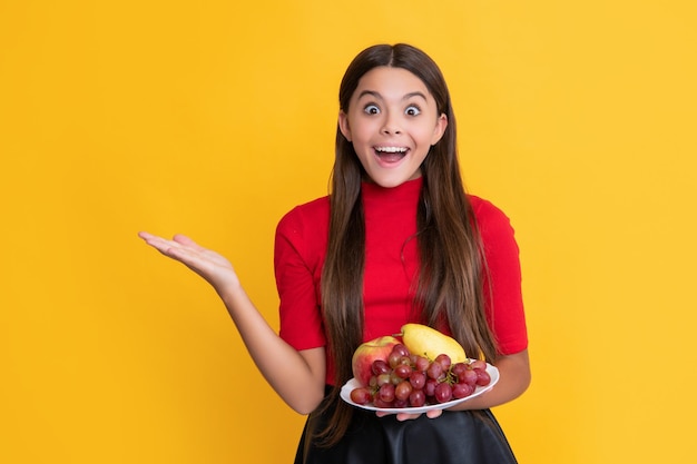 Amazed smiling child hold fresh fruit plate on yellow background