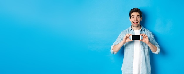 Photo amazed smiling man showing smartphone screen internet promo offer standing against blue background        person