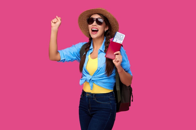 Amazed and smiling woman traveler in straw hat and sunglasses holding plane ticket and passport on pink isolated background