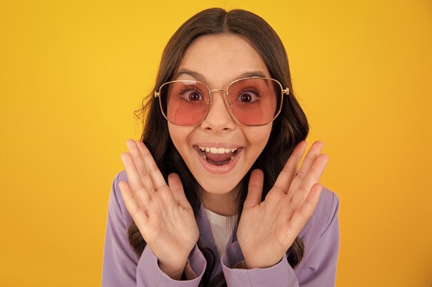 Amazed teen girl excited expression cheerful and glad headshot portrait of cute teenager child girl isolated on yellow studio background wear sunglasses look at camera