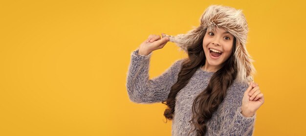 Amazed teen girl wear earflap hat on yellow background keep warm banner of child girl in winter hat studio poster header with copy space