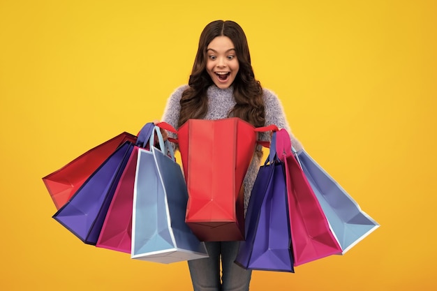 Amazed teenager beautiful fashion teenager child girl with\
shopping bags on yellow background shopaholic shopping and fashion\
kid with shopping sale bags excited teen girl