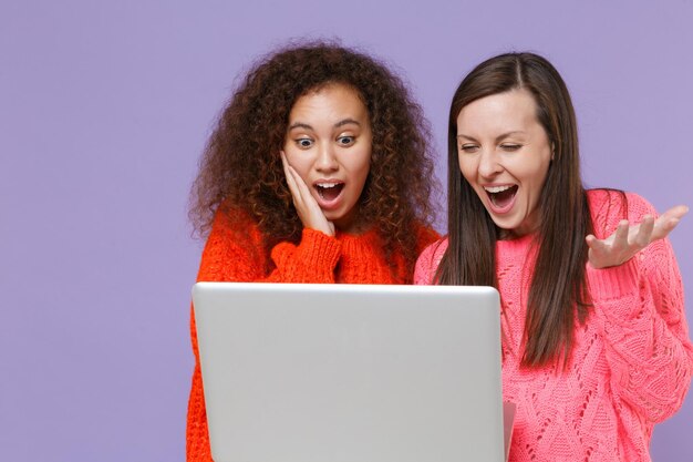 Amazed two young european african american women friends in knitted sweaters isolated on violet purple background studio portrait. people emotions lifestyle concept. working on laptop pc computer