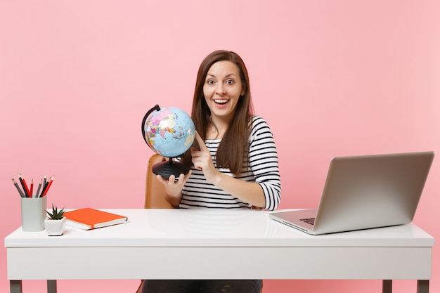Amazed woman holding world globe and planning vacation while sit and work at white desk with contemporary pc laptop isolated on pastel pink background. Achievement business career concept. Copy space.