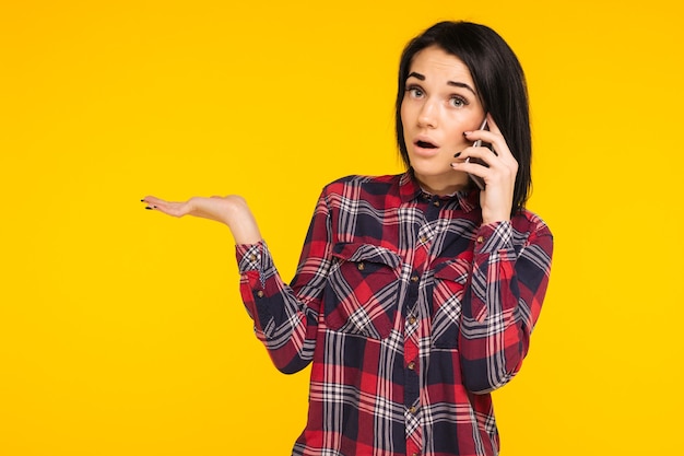 Amazed woman keeping mouth wide open, looking surprised, talking on mobile phone, conducting pleasant conversation isolated on yellow background. People sincere emotions, lifestyle.