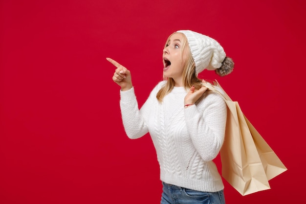 Amazed woman in sweater hat isolated on red background. healthy fashion lifestyle, cold season concept. mock up copy space. hold package bag with purchases after shopping, pointing index finger aside