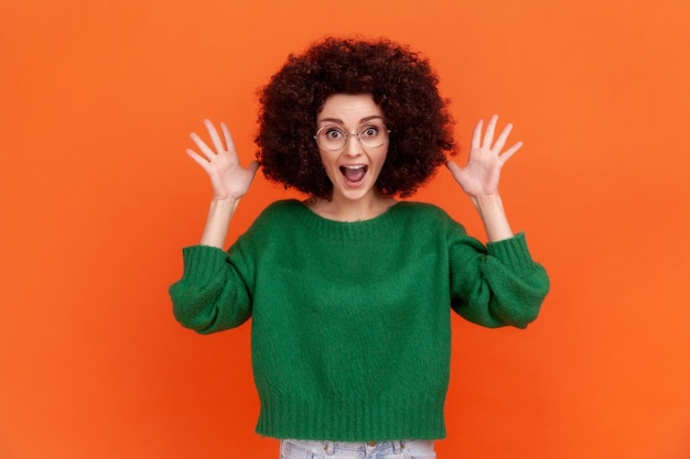 Photo amazed woman with afro hairstyle wearing green casual style sweater and eyeglasses standing with raised arms, yelling with excitement. indoor studio shot isolated on orange background.
