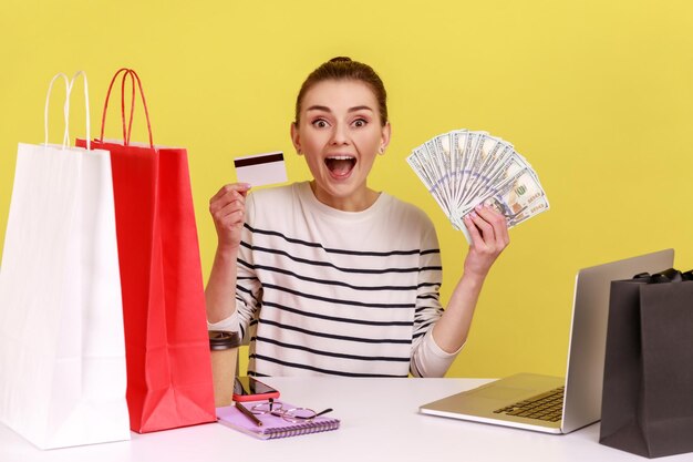 Изумленная женщина на рабочем месте с ноутбуком, держащим доллары и кредитную карту, с улыбкой смотрит в камеру