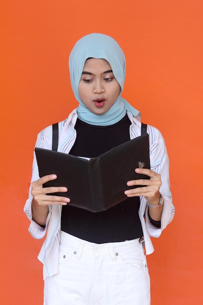 Amazed young Asian girl wearing hijab reading book isolated over orange background