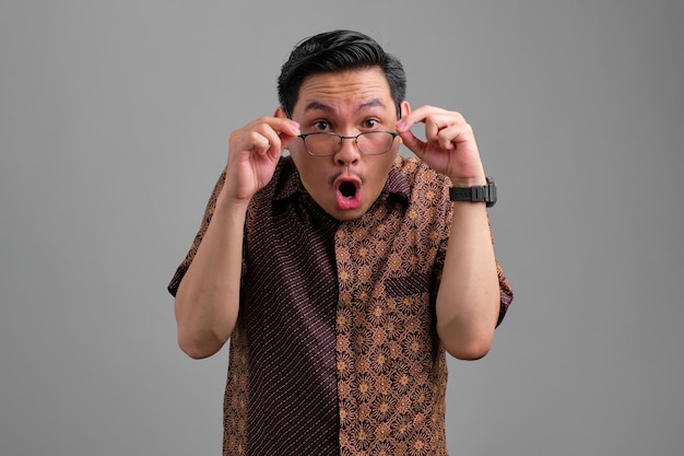 Amazed young Asian man in batik shirt touching glasses with mouth opened reacting to great news isolated on grey background