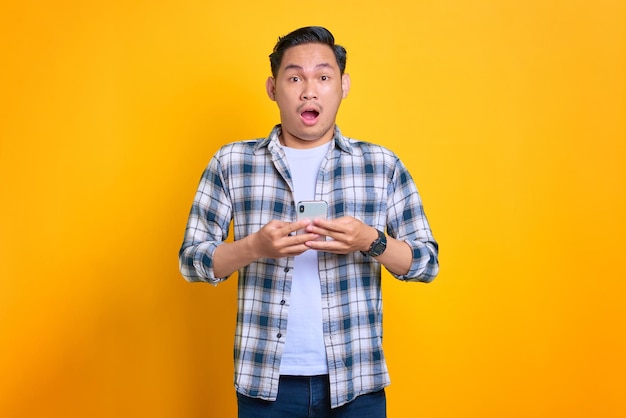 휴대폰을 사용하고 노란색 배경에 격리된 카메라를 보고 있는 격자 무늬 셔츠에 놀란 젊은 아시아 남자