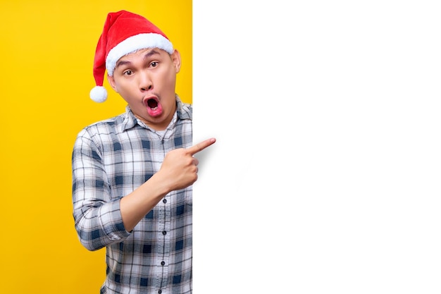 Amazed young asian man wearing christmas hat points out the side of a white advertisement board isolated on yellow background happy new year 2023 holiday concept