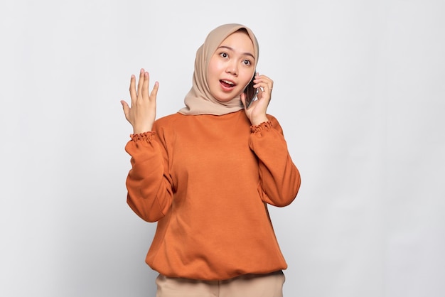 Amazed young Asian woman in orange shirt talking on mobile phone isolated over white background