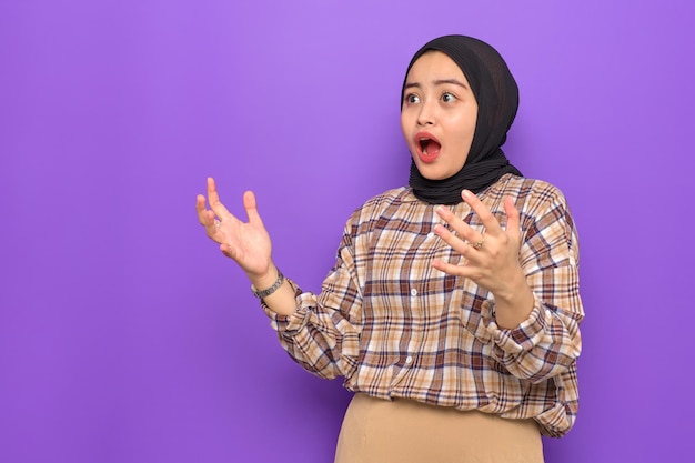 Amazed young asian woman in plaid shirt looking away at copy\
space with wow face expression isolated on purple background