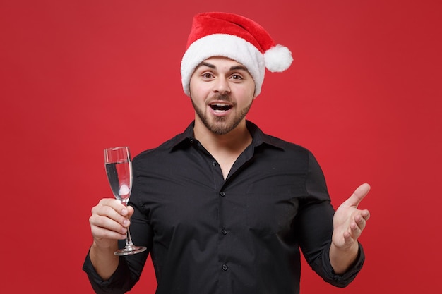 Foto stupito il giovane babbo natale barbuto con la classica camicia nera cappello di natale isolato su sfondo rosso. felice anno nuovo celebrazione concetto di vacanza. mock up spazio di copia. tenere un bicchiere di champagne allargando le mani.