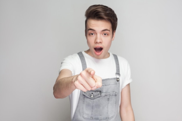 Amazed young brunette man in casual style with t-shirt and denim overalls standing, looking and pointing at camera with surprised unbelievable face. indoor studio shot, isolated on gray background.