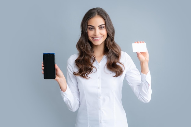 Amazed young brunette woman in shirt using mobile cell smart phone hold credit bank card isolated