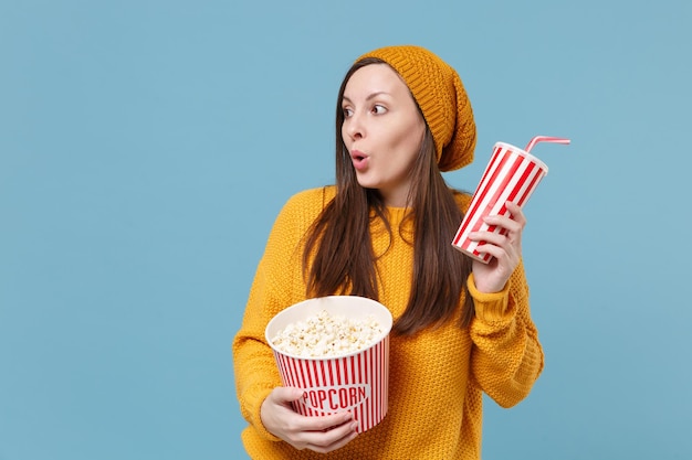 Amazed young brunette woman in sweater hat posing isolated on\
blue background. people emotions in cinema lifestyle concept. mock\
up copy space. watching movie film hold bucket of popcorn cup of\
soda.