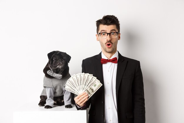 Amazed young man in party suit, standing near cute black pug dog in costume