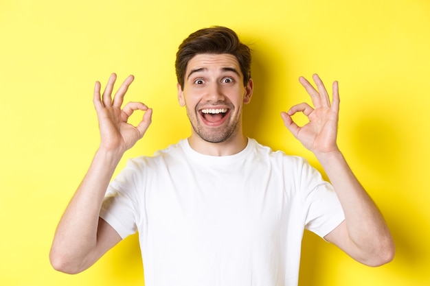 Amazed young man showing ok signs and smiling, recommending something good, standing over yellow wall