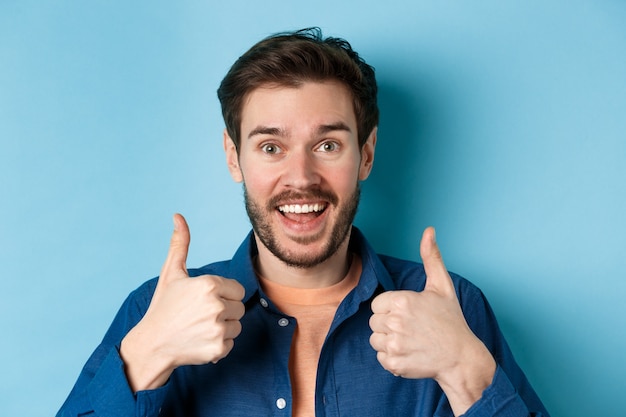 Amazed young man smiling and showing thumbs up in approval, like something good, praise excellent choice, standing on blue background.