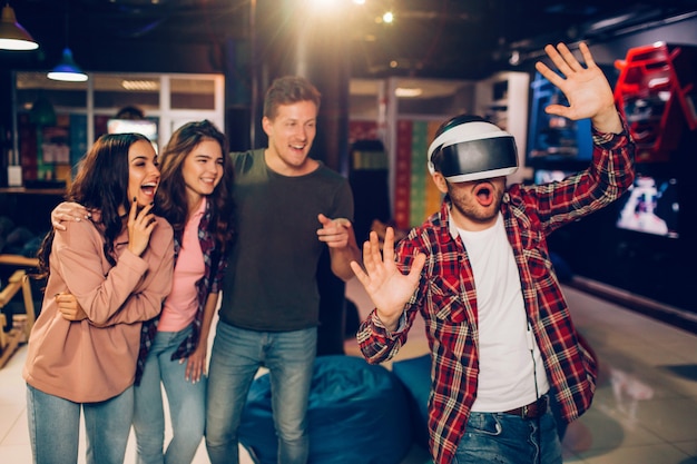 Amazed young man wearing VR glasses and wave hands in playing room. His friends look happy and excited. They stand behind.