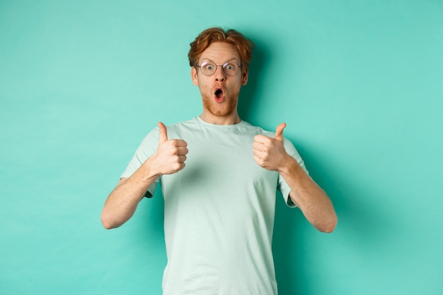 Amazed young man with red hair and beard, wearing glasses with t-shirt, showing thumbs-up and gasping in awe, checking out awesome promo offer, turquoise background