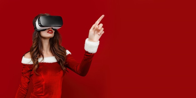 Amazed young santa girl in christmas dress looking in headset pointing hands aside isolated on red w...