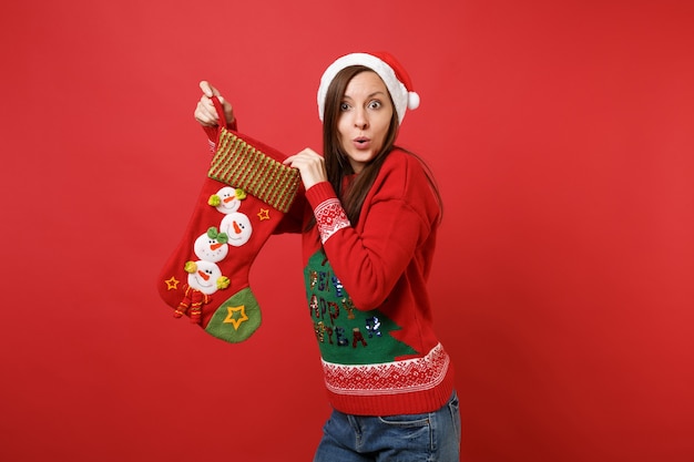 赤い壁の背景に隔離された贈り物のためのストッキング、靴下を持って、驚いたように見えるクリスマス帽子の驚いた若いサンタの女の子。明けましておめでとうございます2019お祝いホリデーパーティーのコンセプト。コピースペースをモックアップします。