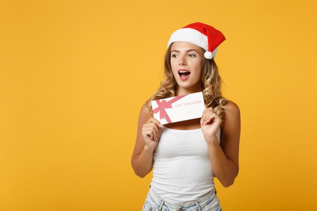 흰 셔츠를 입은 어린 산타 소녀, 스튜디오의 노란색 배경에 고립된 포즈를 취한 크리스마스 모자에 놀랐습니다. 새해 복 많이 받으세요 2020 축 하 휴일 개념입니다. 복사 공간을 비웃습니다. 상품권을 들고.