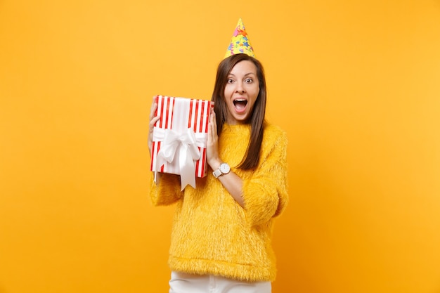 La giovane donna stupita in cappello della festa di compleanno tiene la scatola rossa con il regalo presente che celebra godendo la festa isolata su fondo giallo luminoso. persone sincere emozioni, concetto di stile di vita. zona pubblicità.