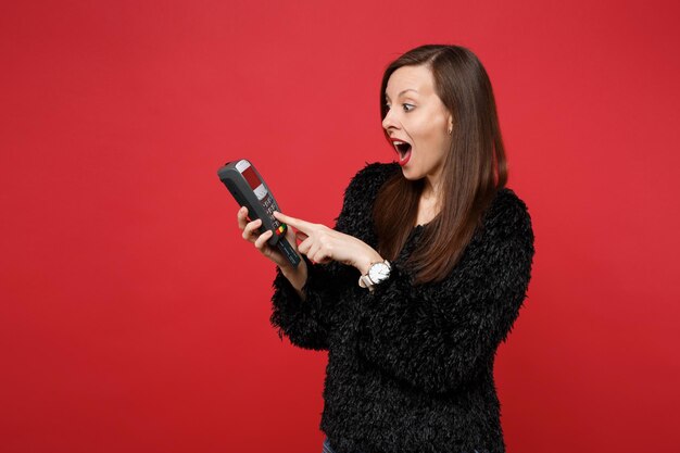 Пораженная молодая женщина в черном меховом свитере держит беспроводной современный банковский платежный терминал для обработки и получения платежей по кредитным картам, изолированных на красном фоне. Концепция образа жизни искренние эмоции людей.