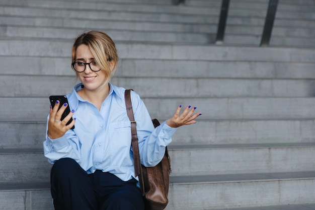 Пораженная молодая женщина в очках сидит на ступеньках и смотрит на экран смартфона. Красивая женщина в голубой рубашке позирует с современным устройством на открытом воздухе.