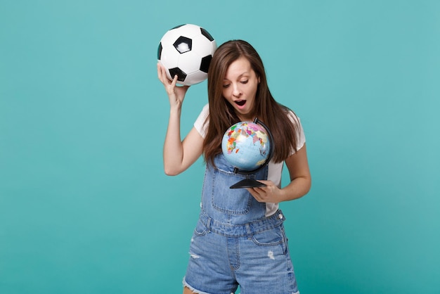 Фото Изумленная молодая женщина-футболист держит футбольный мяч, глядя на земной шар, изолированный на синем бирюзовом фоне. эмоции людей, концепция спортивного семейного отдыха. скопируйте пространство для копирования.