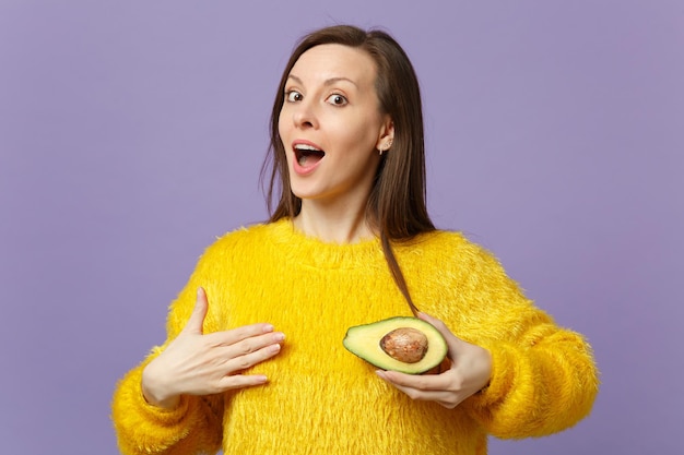 Amazed young woman in fur sweater keeping mouth open holding half of fresh ripe green avocado isolated on violet pastel background. People vivid lifestyle, relax vacation concept. Mock up copy space.