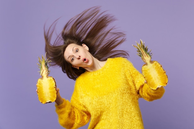 Фото Изумленная молодая женщина в меховом свитере с развевающимися волосами держит половинки свежих спелых ананасов, изолированных на фиолетовом пастельном фоне. люди яркий образ жизни расслабляют отпуск концепция. скопируйте пространство для копирования.
