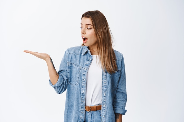 Amazed young woman looking at her arm as if holding something beautiful in palm against empty copy space, standing in casual clothes and advertising product, white wall