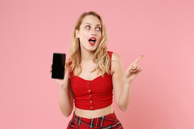 Amazed young woman in red sexy clothes posing isolated on pastel pink background. People lifestyle concept. Mock up copy space. Hold mobile phone with blank empty screen pointing index finger aside.