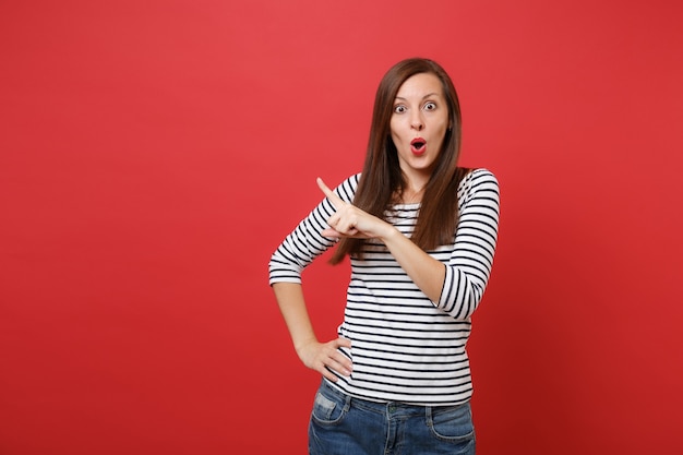 Amazed young woman in striped clothes pointing index finger aside keeping mouth wide open, looking surprised 