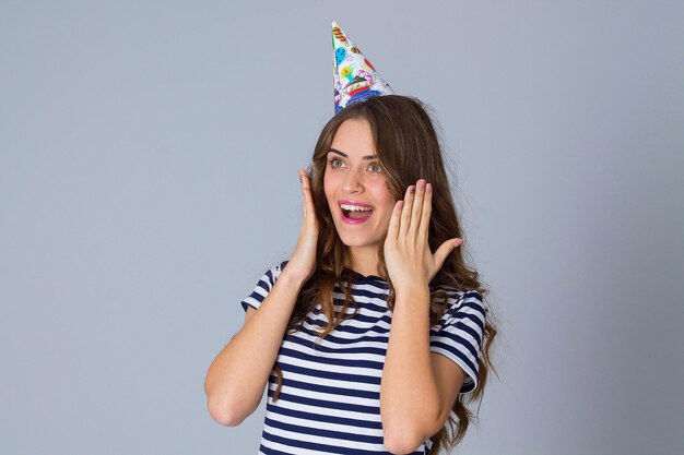 Amazed young woman wearing in stripped T-shirt and celebration cap on grey background in studio