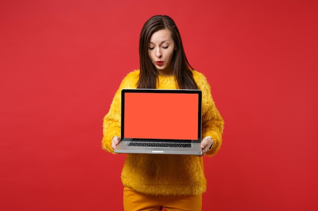 Giovane donna stupita in maglione di pelliccia gialla che guarda sul computer del pc portatile con schermo vuoto vuoto nelle mani isolate sul fondo rosso della parete. persone sincere emozioni, concetto di stile di vita. mock up copia spazio.