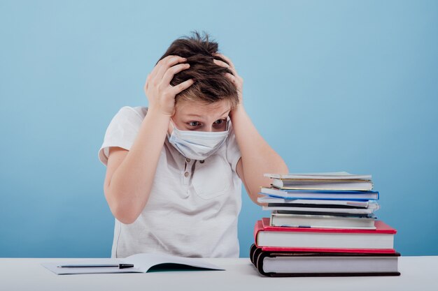 Amazement boy with medical mask looking at books sitting at the table isolated on blue background