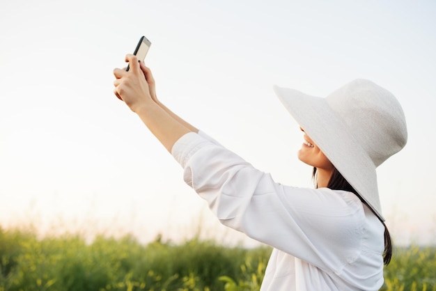 自然フィールドの背景Instagramの電話で笑顔とselfieを取る帽子と白いシャツを持つ驚くべき魅力的なロマンチックな若い女性