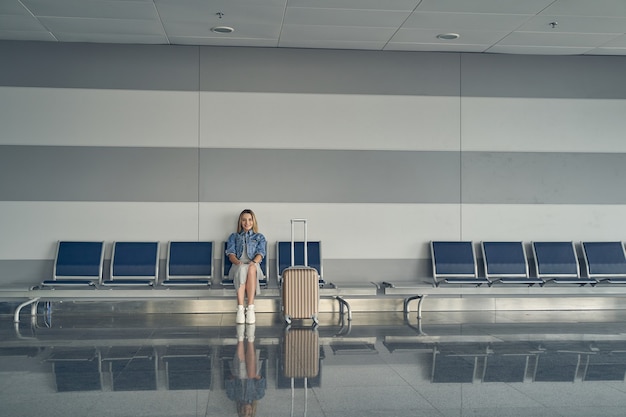 Foto incredibile donna bionda che esprime positività mentre aspetta il suo volo, sognando una vacanza attiva