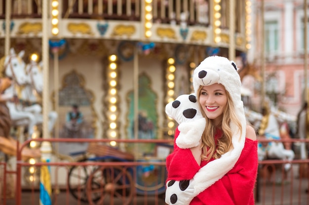 Amazing blonde woman wearing red knitted sweater and funny hat, posing at the background of carousel with lights