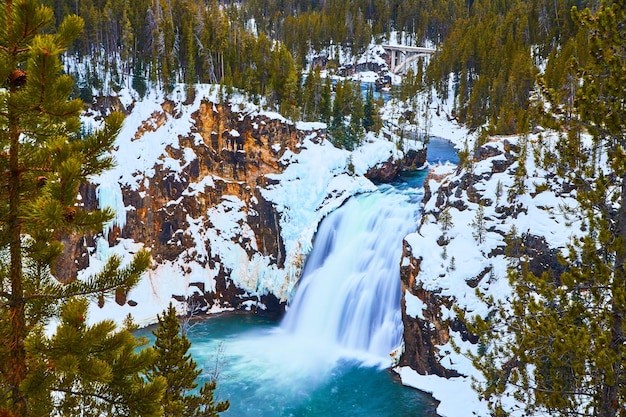 Amazing blue and peaceful waterfall by snowy cliffs and pine trees in Yellowstone