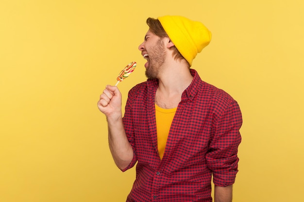 Amazing carefree hipster guy in beanie hat, checkered shirt\
screaming into lollipop like microphone, singing loudly as popstar\
in karaoke, having fun. indoor studio shot isolated on yellow\
background