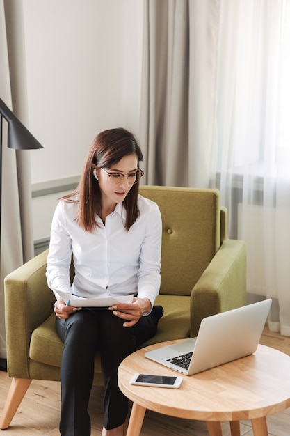 amazing concentrated beautiful young business woman in formal wear clothes indoors at home talking using laptop computer with earphones holding documents.